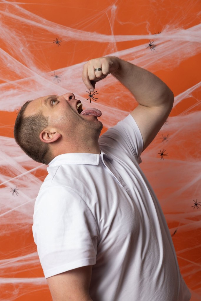 A man playfully pretending to eat a fake spider while making a surprised expression, set against an orange background with spider web decorations.