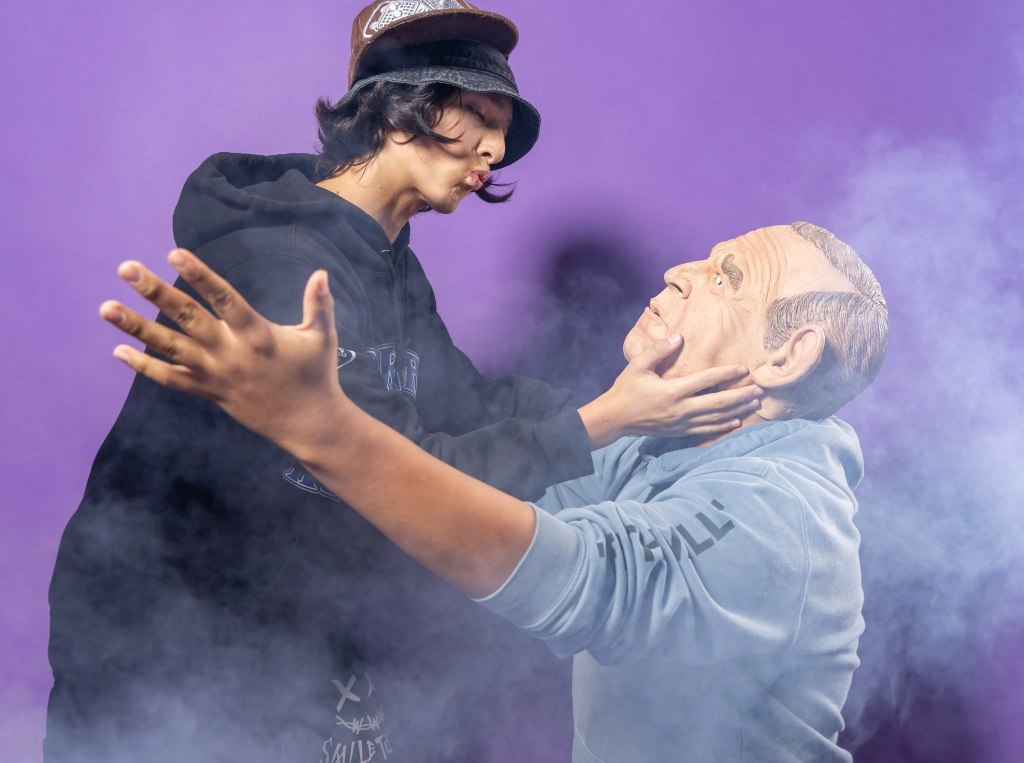 A person wearing a black hoodie and hat leans in to kiss a lifelike statue of a man, set against a purple background with wisps of smoke.