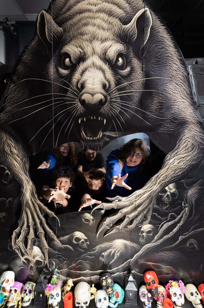 A group of four people poses in front of a large, fearsome illustration of a rat and skeletal hands, with colorful skull decorations and candles in the foreground.
