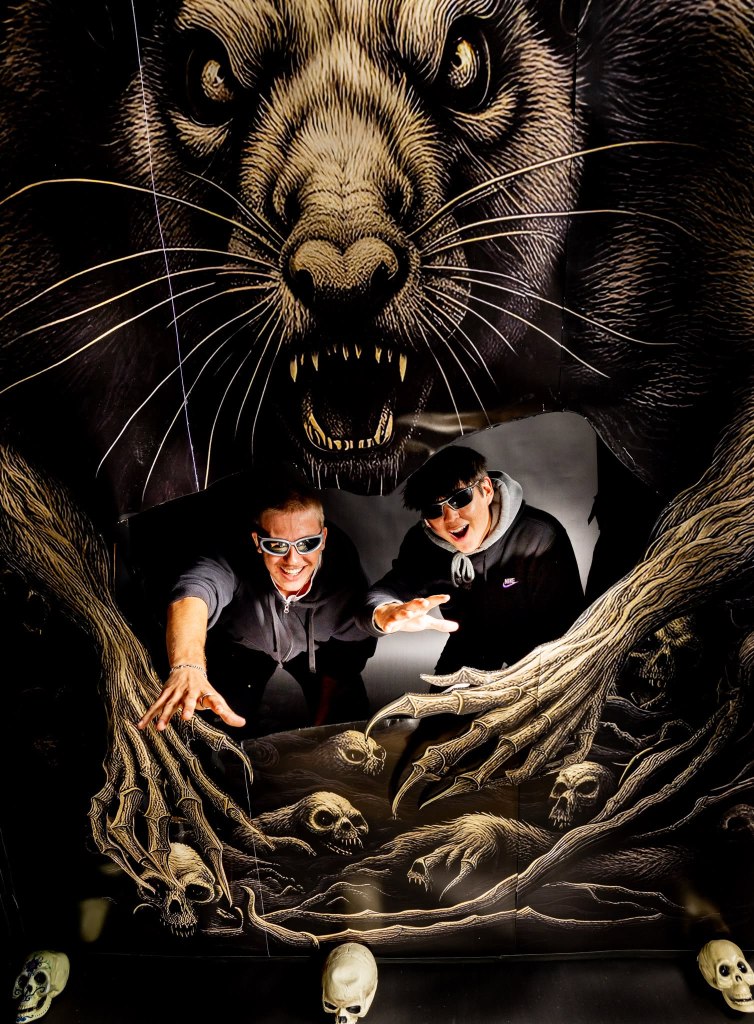 Two individuals posing playfully in front of a large, dramatic mural featuring a menacing rat with bared teeth and clawed hands reaching toward skulls and other eerie elements.
