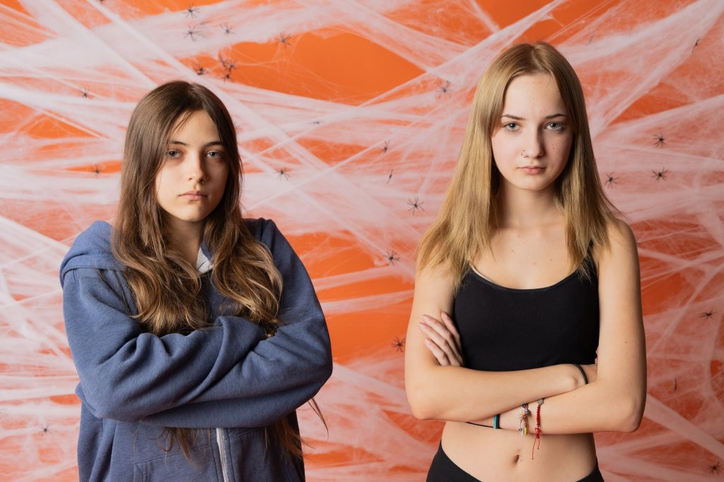 Two teenage girls standing side by side with serious expressions, one in a blue hoodie and the other in a black tank top, against an orange background decorated with spider webs and spiders.