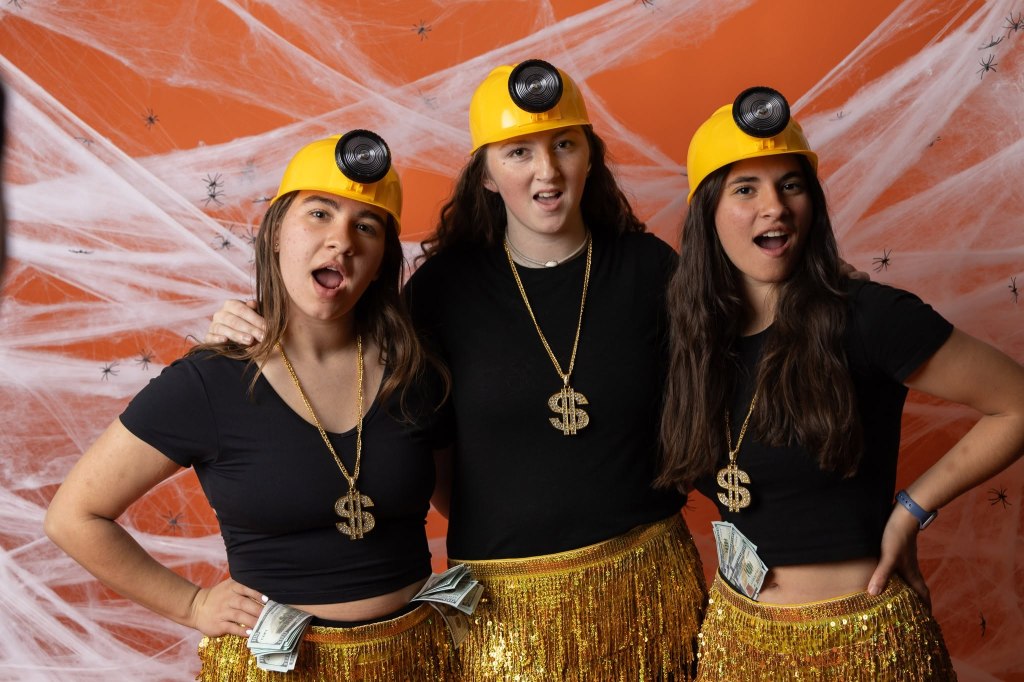 Three women posing in fun costumes with yellow miner hats and gold sequined skirts, accessorized with dollar sign necklaces and fake money. Background features Halloween-themed spiderwebs.