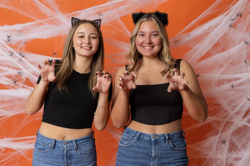 Two young women in black tops and jeans pose playfully with clawed hands, wearing cat ear headbands, against an orange backdrop decorated with spider web and plastic spiders.