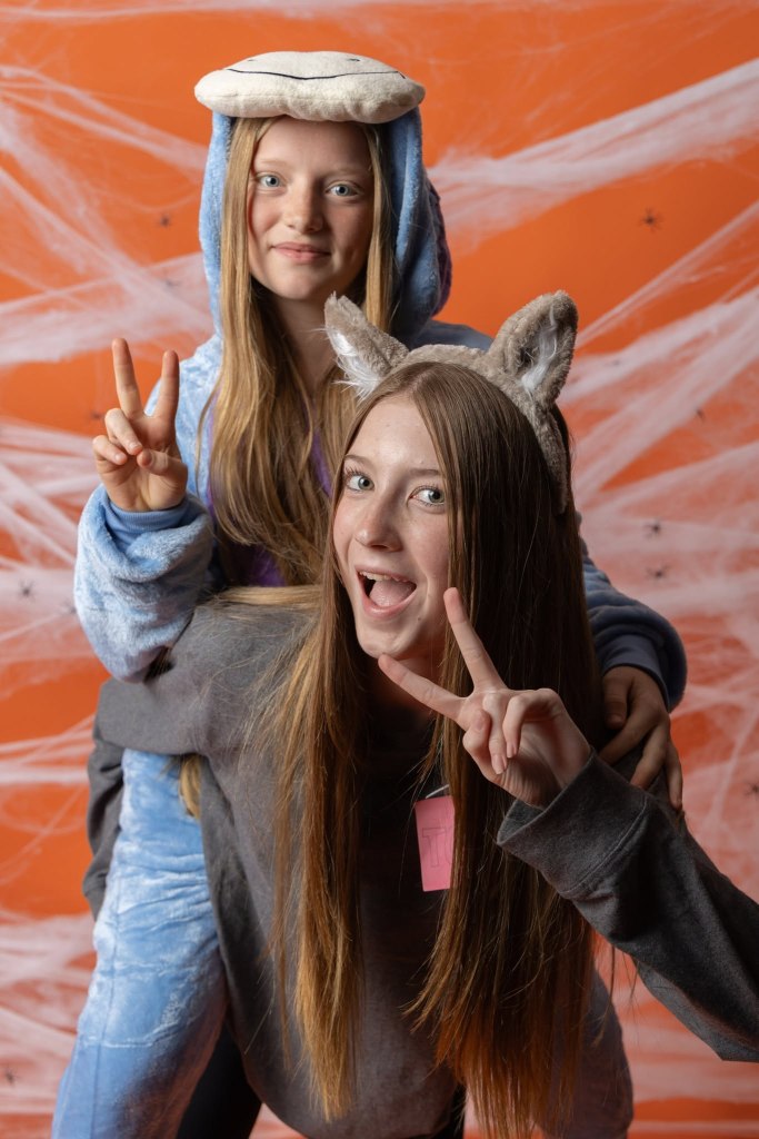 Two girls posing for a fun photo; one wearing a blue animal onesie and the other in a gray sweatshirt with cat ears. They are both making peace signs and smiling against an orange background decorated with spider webs.