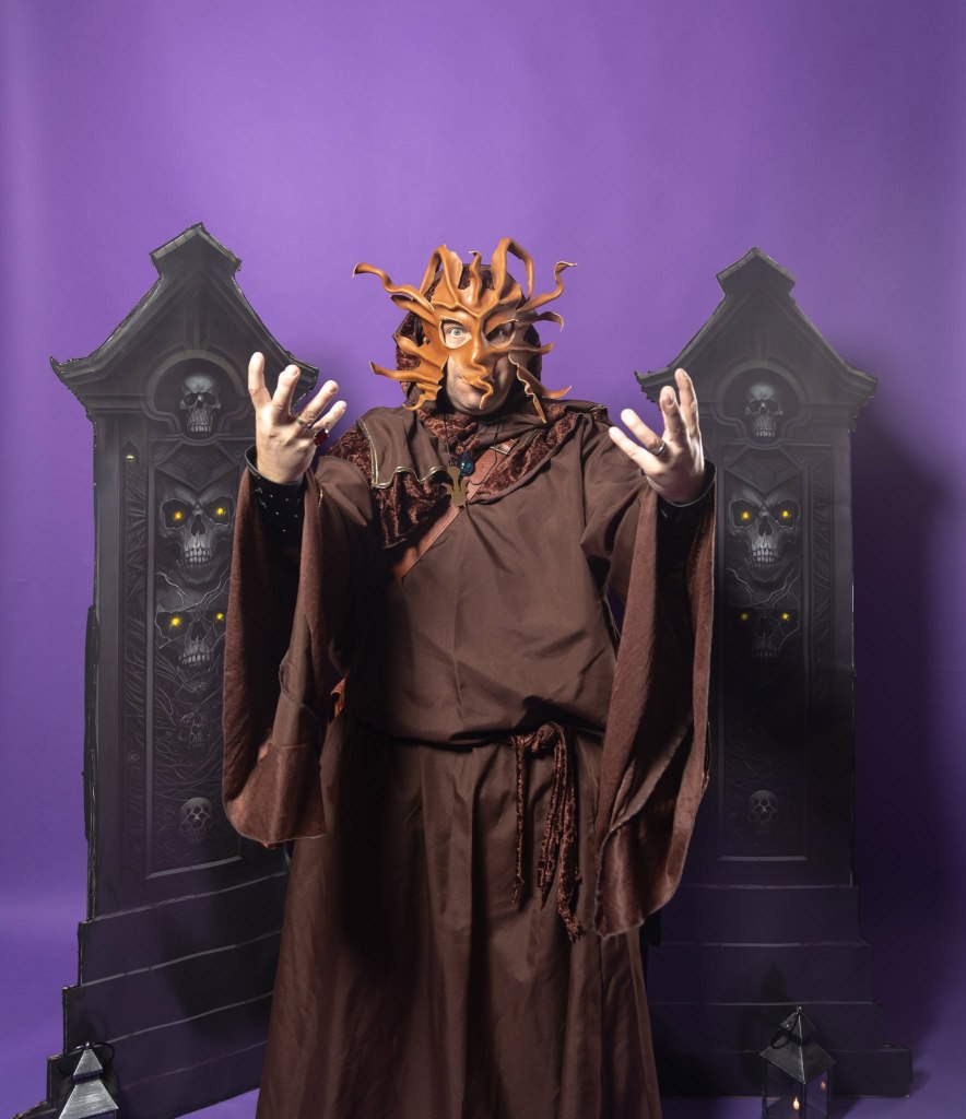 Person in a brown robe and an elaborate mask resembling tree branches, posing dramatically against a purple backdrop with gothic-style decorations featuring skulls and glowing eyes.