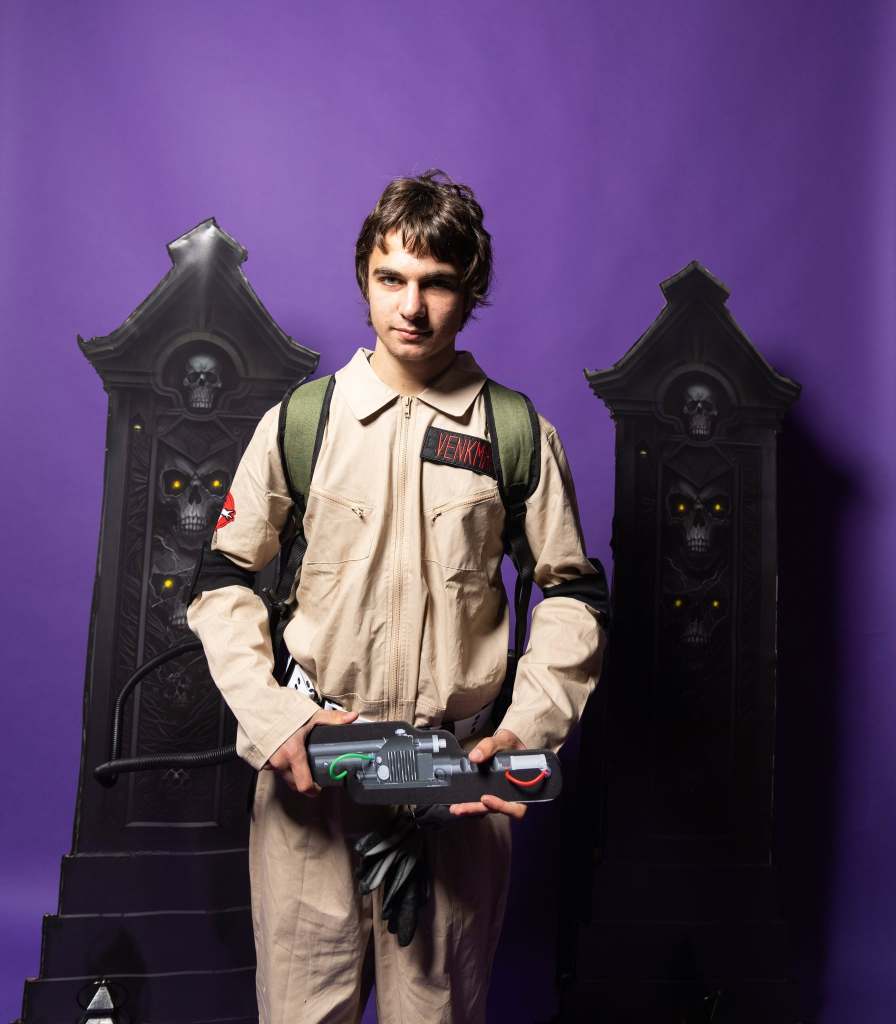 A young man wearing a beige Ghostbusters uniform with a green backpack, holding a ghost-catching device, standing in front of spooky black decor on a purple background.