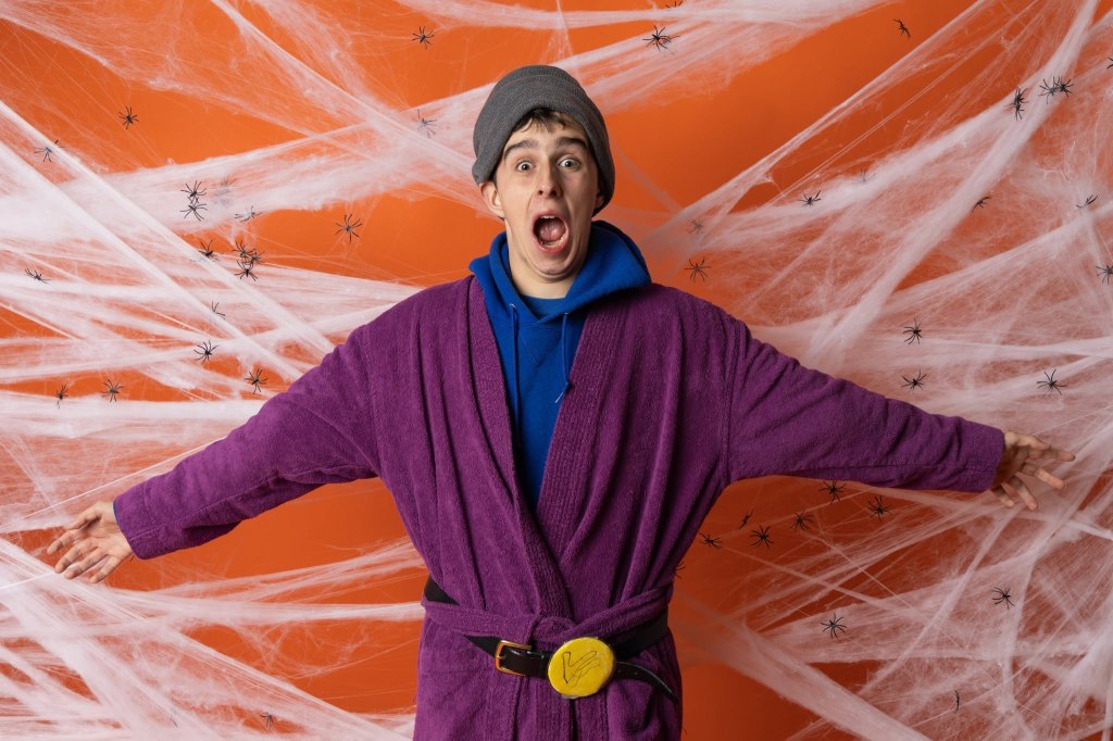 A person wearing a purple robe and a gray beanie stands with arms outstretched against an orange background adorned with spider webs and plastic spiders.