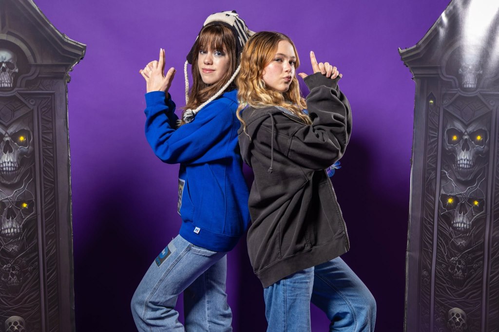 Two individuals pose back-to-back against a purple backdrop, one wearing a blue hoodie and the other in a black hoodie, both making playful hand gestures. They are flanked by large, decorative panels featuring skull designs.