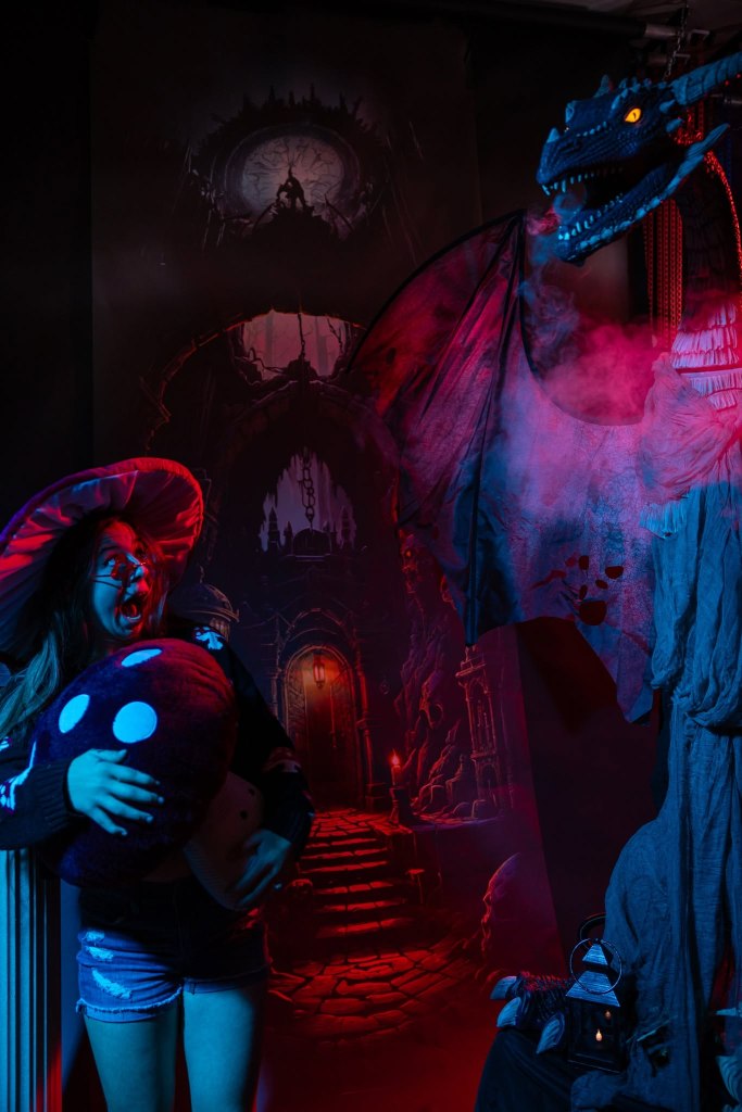 A woman wearing a large red hat and holding a plush mushroom toy stands in front of a dramatic fantasy backdrop featuring a dragon, mysterious fog, and a shadowy pathway.