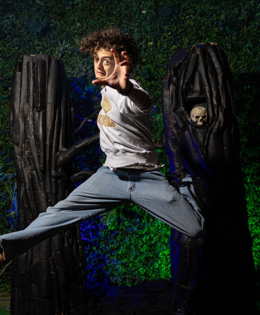 A person with curly hair is jumping in front of two large black tree-like structures, with a skull partially visible in one of them, surrounded by greenery and colorful lighting.