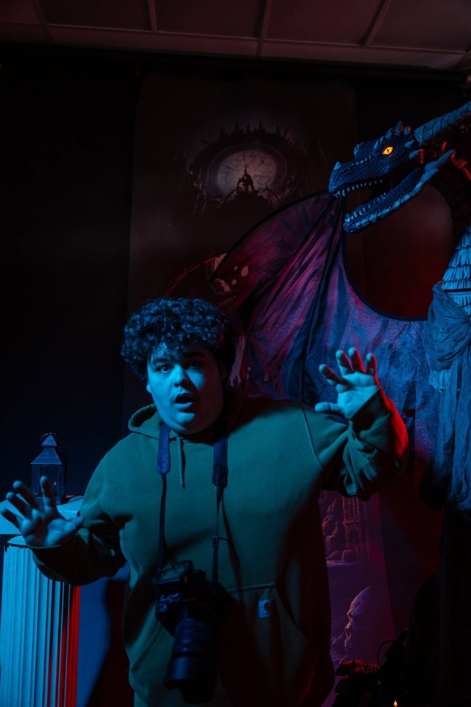 A surprised young man in a green hoodie with a camera around his neck appears startled in front of a menacing dragon statue, set against a dark, atmospheric backdrop.