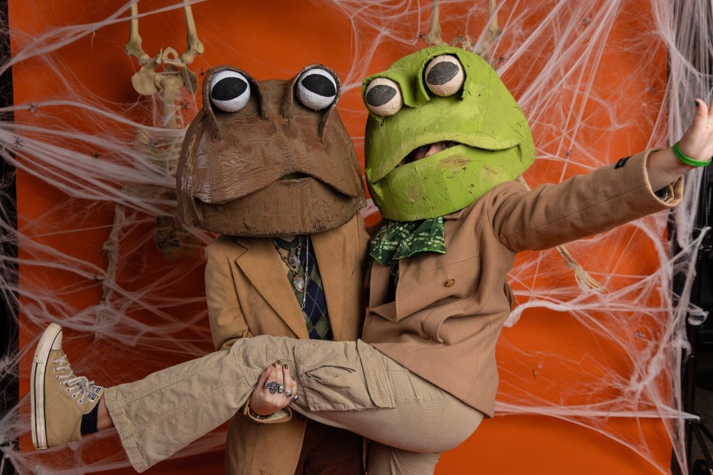 Two individuals wearing oversized frog masks, one brown and one green, in Halloween-themed attire. They are posing playfully in front of an orange background with spider web decorations.