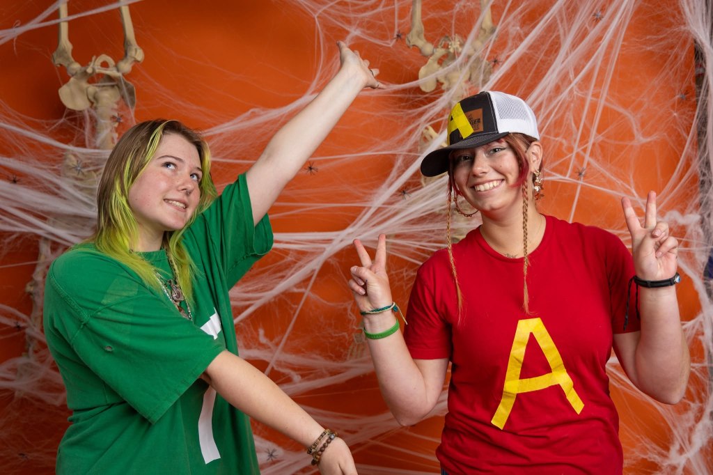 Two young women posing in front of an orange wall decorated with spider webs and skeletons. One is wearing a green shirt and pointing upwards, while the other is wearing a red shirt and peace-signing.