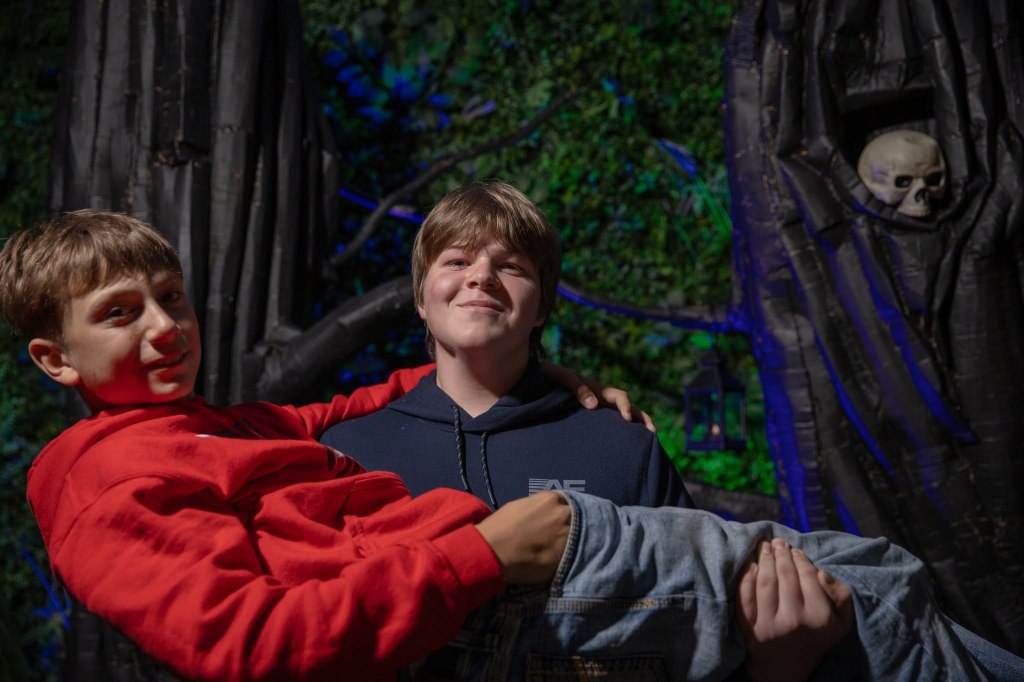Two young boys posing in a playful manner, with one boy holding the other in his arms against a backdrop of dark trees and a skull decoration.