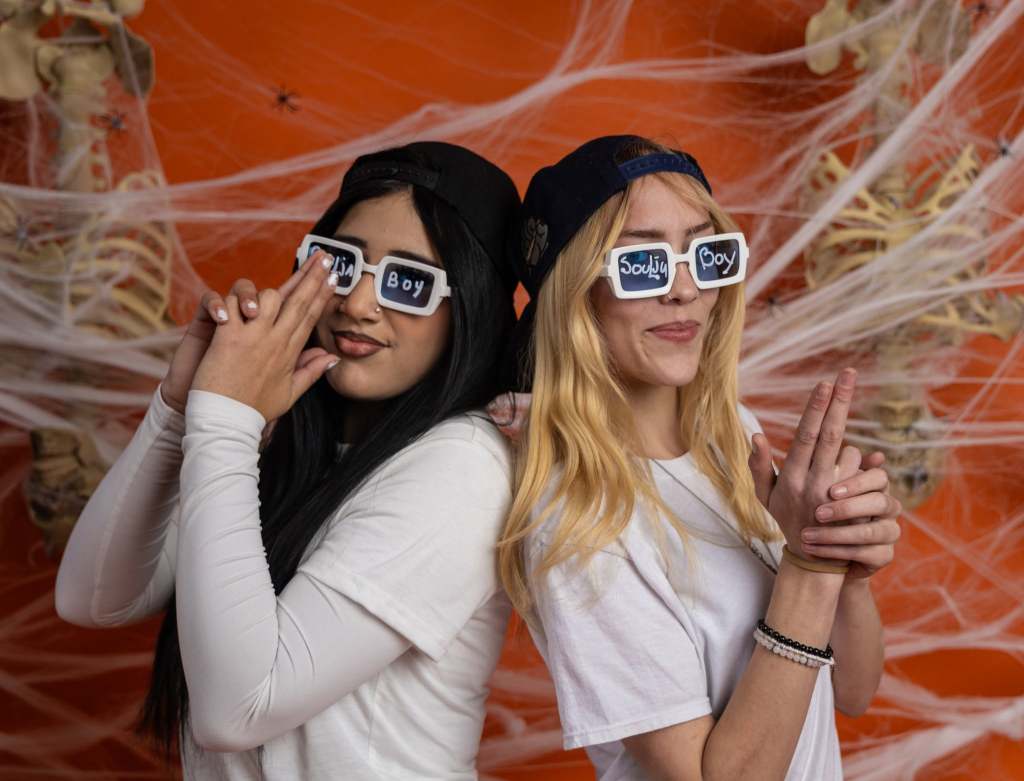 Two women pose playfully against an orange background with spiderweb decorations. They are wearing white t-shirts and sunglasses with the phrases 'Soulja Boy' and 'Dria Boy'.