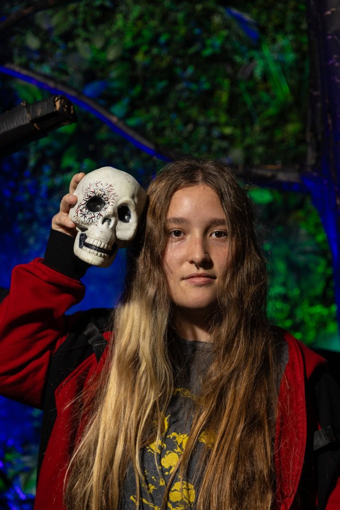 A young person with long hair holding a decorative skull against a vibrant green and blue background.