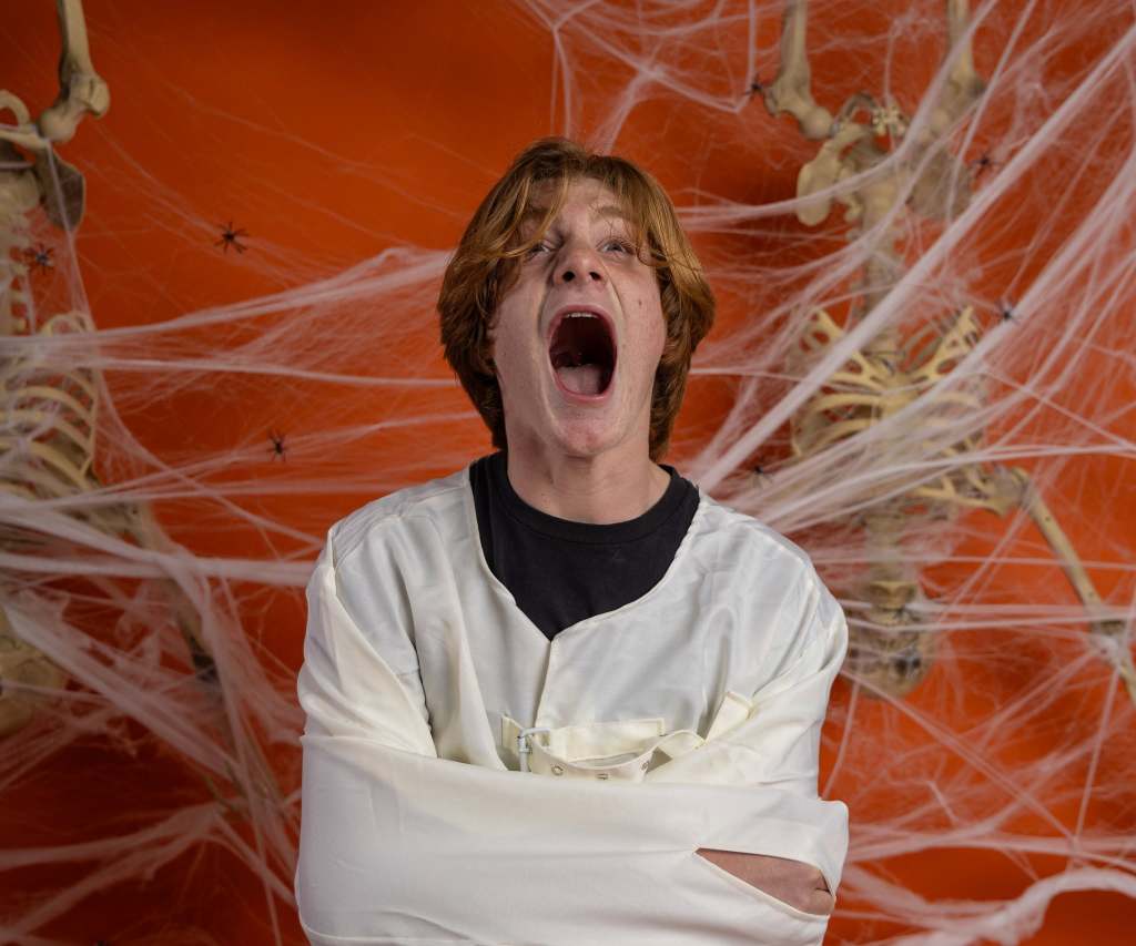 A young man in a straitjacket yelling against an orange background decorated with spiderwebs and skeletons.