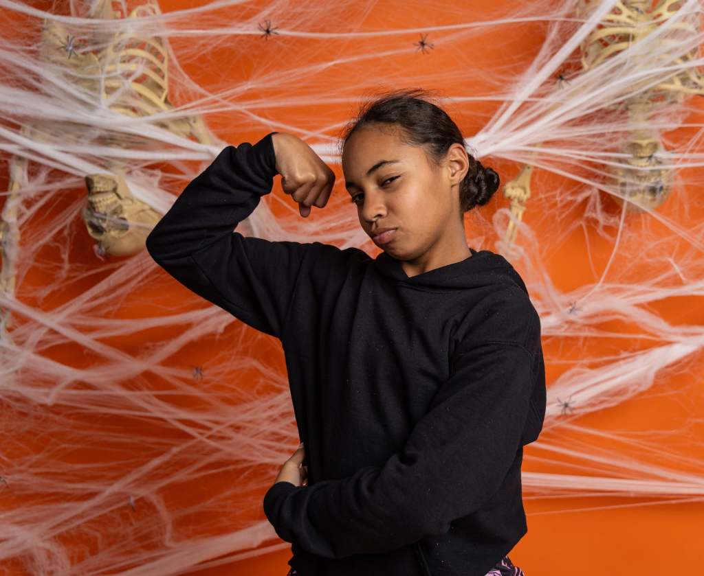 A young girl flexes her arm muscle playfully in front of an orange background decorated with Halloween-themed spider webs and a skeleton.
