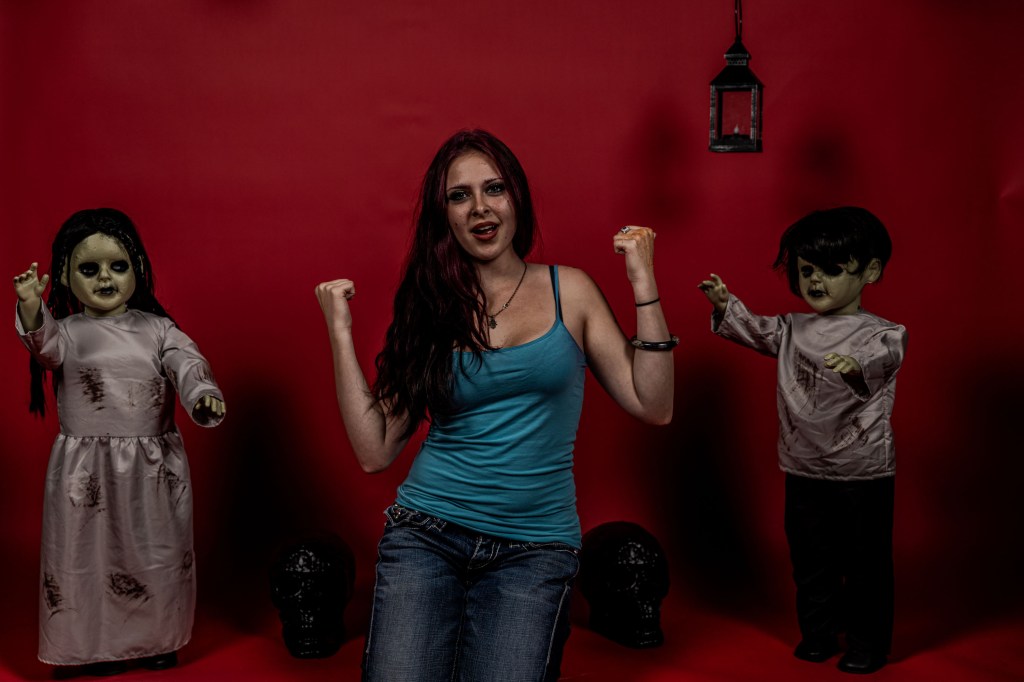 A woman in a blue tank top poses in front of a red background, surrounded by two eerie dolls, one resembling a zombie and the other a ghost-like figure. The setting includes decorative skulls and a lantern hanging above.