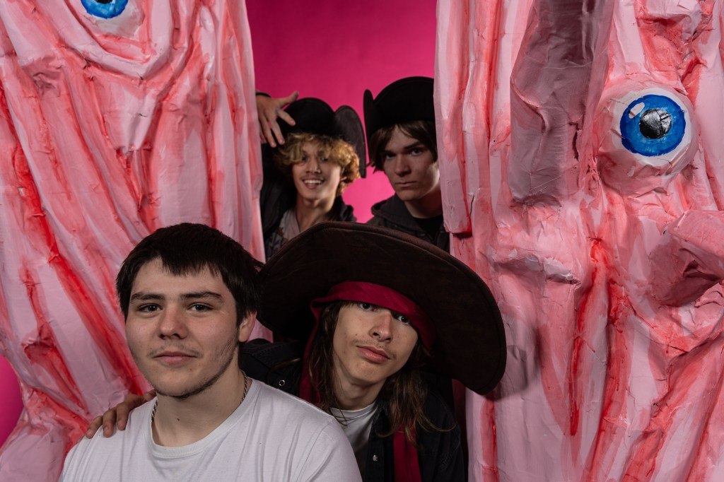 A group of four individuals posing in front of large, pink, monster-like props with exaggerated features and a vibrant pink background.