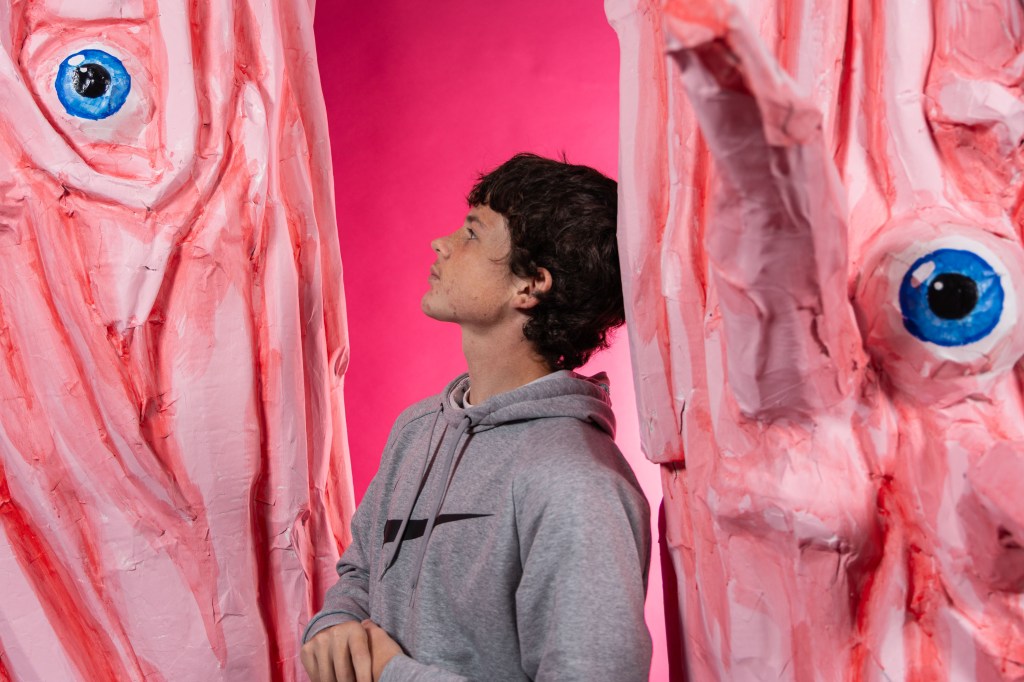 A young man in a gray hoodie stands between two large, abstract pink paper sculptures with exaggerated blue eyes, against a pink background.