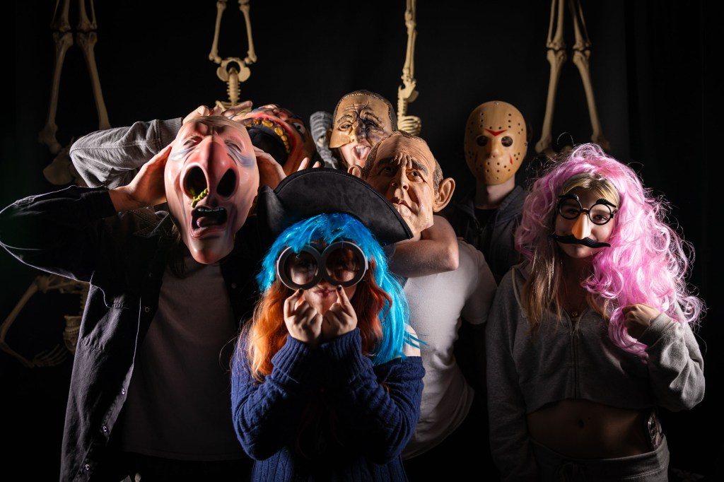 A group of people wearing various theatrical and horror masks, posing against a dark background with skeleton decorations.