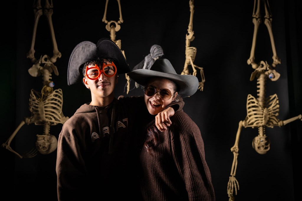 Two individuals posing playfully in front of a black backdrop with skeleton decorations, one wearing oversized orange glasses and a pirate hat, the other in a large grey hat and sunglasses.