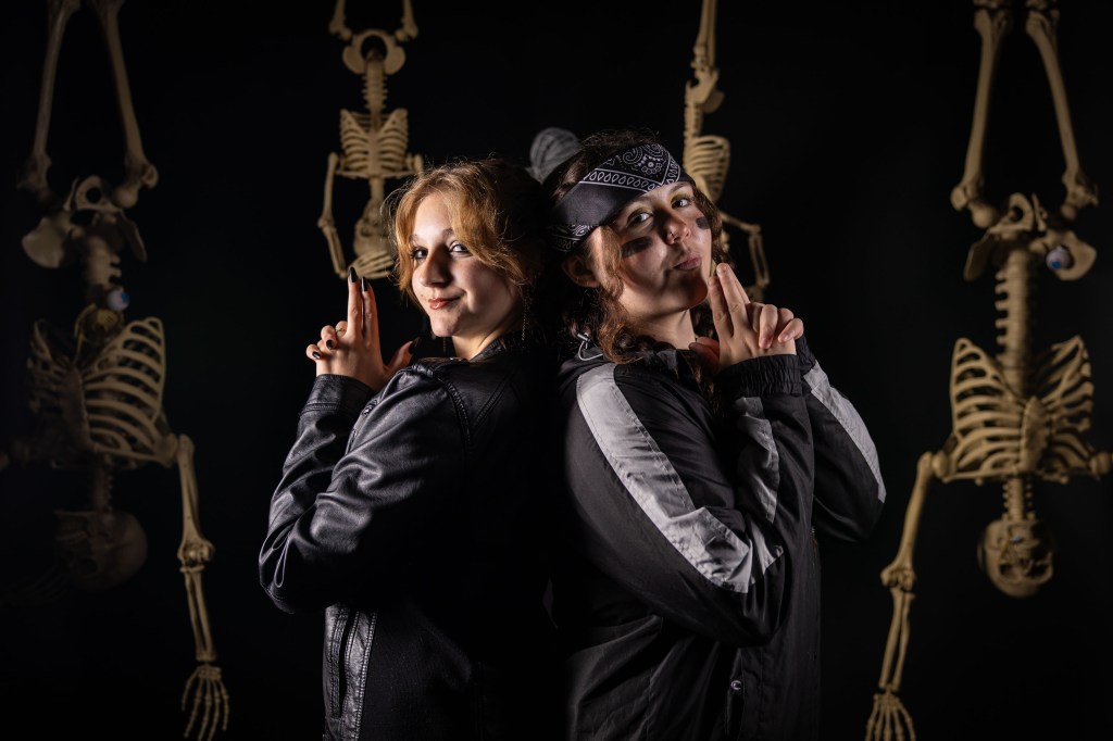Two individuals posing back-to-back with playful expressions, set against a backdrop of hanging skeletons in a dark environment.