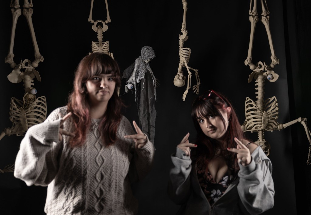 Two young women posing in front of a black background decorated with hanging skeletons. One woman is making a peace sign, while the other is gesturing with her hands in a playful manner.