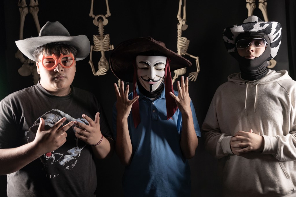 Three individuals dressed in playful costumes and accessories, posing for a photo with skeletons in the background. The first person wears a cowboy hat and oversized glasses, the second wears a Guy Fawkes mask and a hat, and the third wears a patterned hat and hoodie.