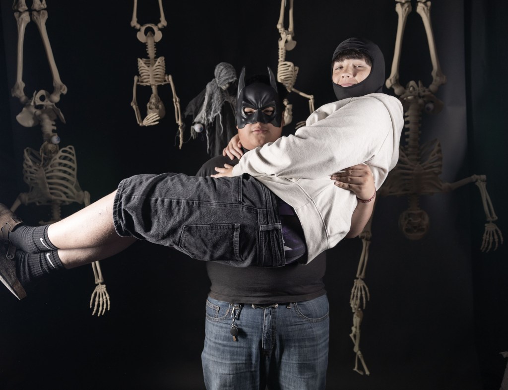 A person in a bat-themed mask holds another individual in a light hoodie, set against a backdrop of hanging skeletons.