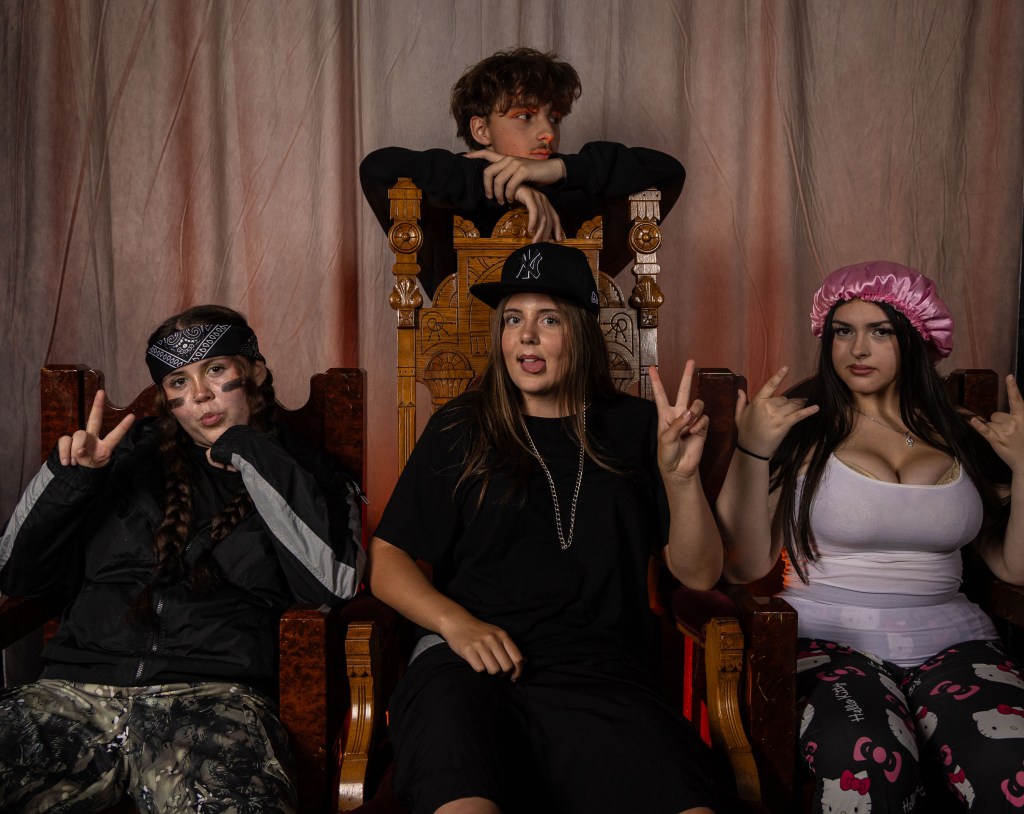 A group of four young people posing together in a playful manner. Two girls are sitting on carved wooden chairs, both making peace signs with their fingers. The girl on the left wears a bandana and camo pants, while the girl in the middle has long hair and wears a black outfit. A third girl sits beside her, wearing a pink shower cap and showing a rock sign. In the background, a boy lounges on a throne-like chair, looking at the camera.