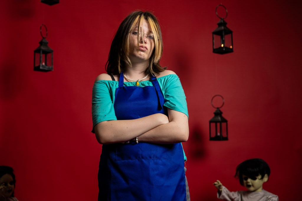A young person with shoulder-length hair standing with arms crossed, wearing a blue apron over a teal shirt. The background is a bright red, featuring hanging lanterns and eerie dolls in the foreground.