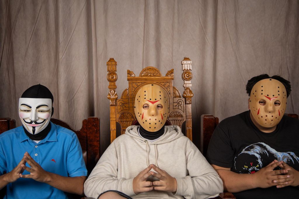 Three individuals wearing distinct masks sit in front of a wooden throne, with a fabric backdrop. The person on the left is wearing a Guy Fawkes mask, the middle person has a hockey mask, and the person on the right also has a hockey mask.