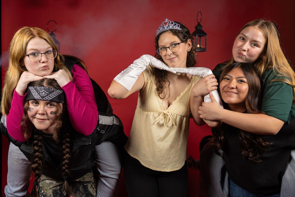 A group of five young women posing playfully together in front of a red background. One woman is on another's back, while the others are standing close together, making fun expressions and showcasing a mix of casual and dressy attire.
