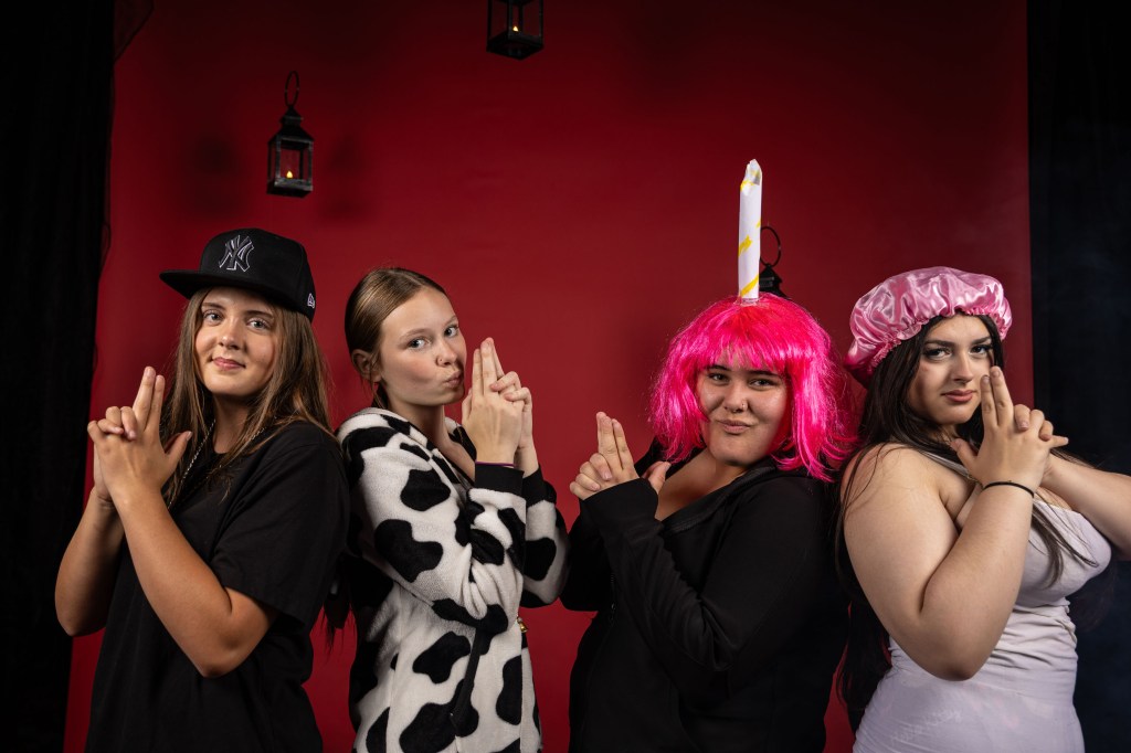 Four individuals posing playfully against a red backdrop, each making gun shapes with their hands. They wear a variety of outfits, including a cow-patterned sweater, a black hoodie, and colorful wigs.