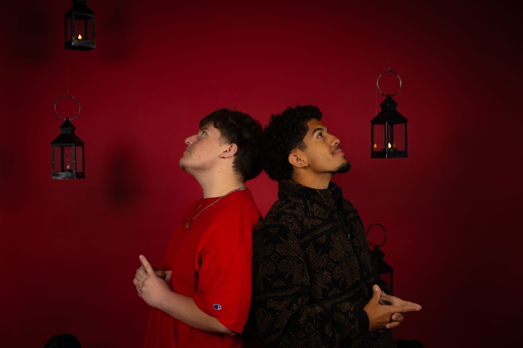 Two young men pose back to back against a red backdrop, looking upwards. Hanging lanterns provide soft lighting in the background.