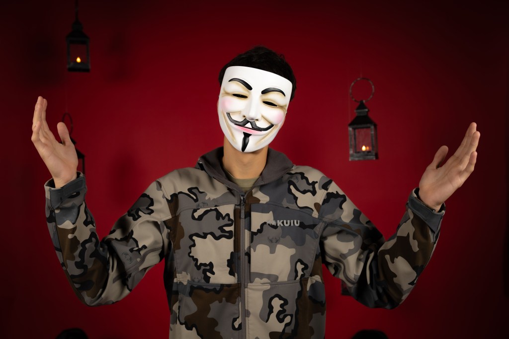 A person wearing a Guy Fawkes mask stands with arms raised in a camouflaged jacket against a red background, with illuminated lanterns in the background.