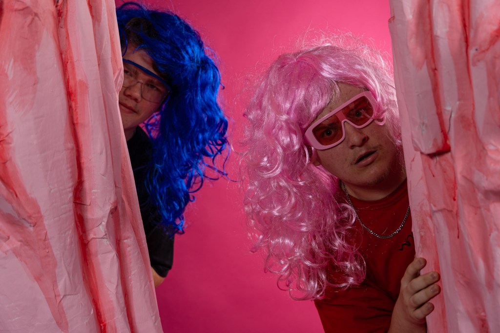 Two individuals with colorful wigs, one blue and one pink, wearing oversized sunglasses, peeking from behind pink fabric against a bright pink background.