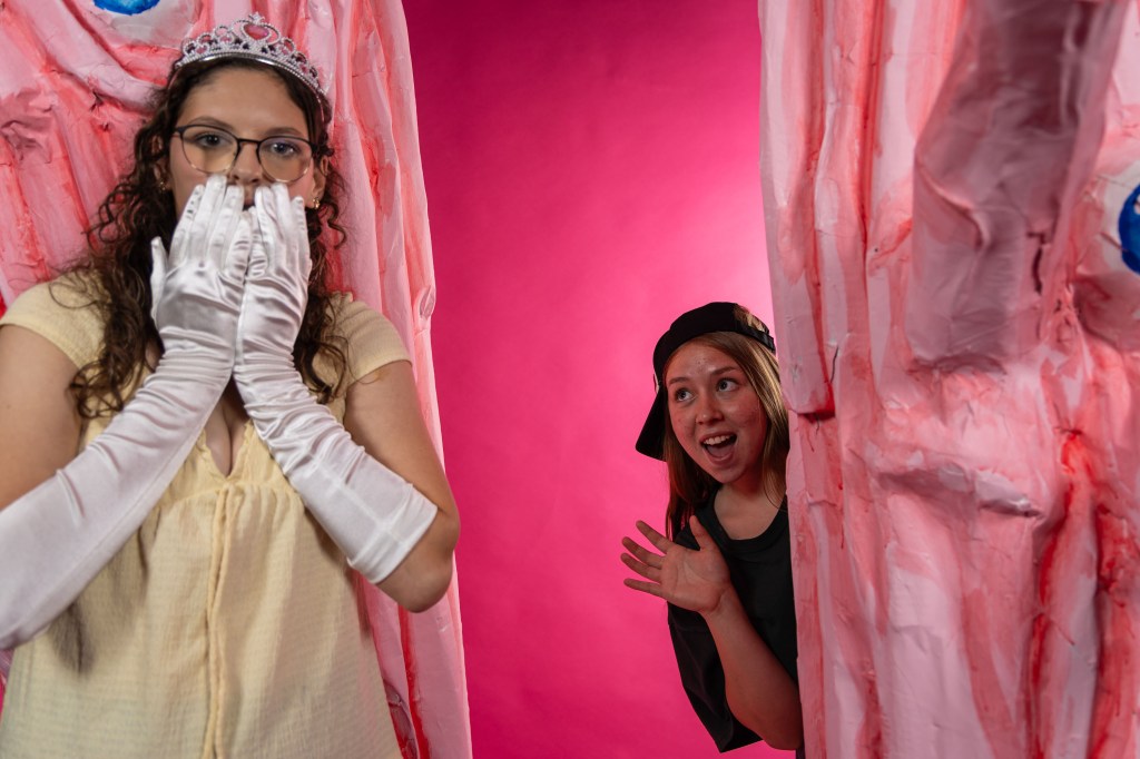 A surprised woman in a yellow dress and white gloves stands against a vibrant pink background, while another woman in a black hat peeks out playfully from behind a large, pink, artistic structure.