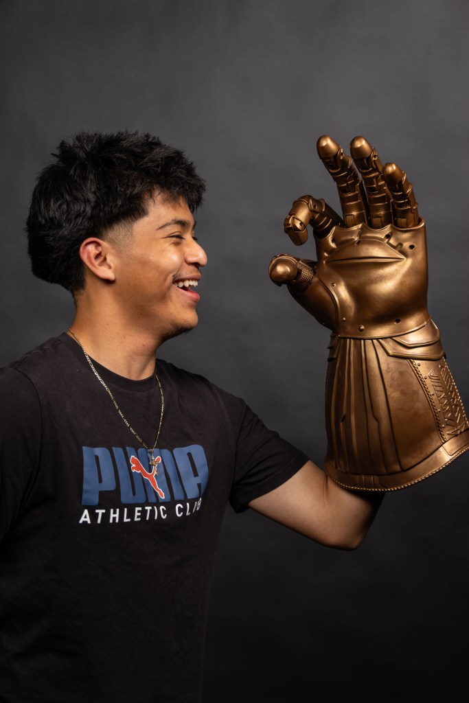 A young man with short dark hair smiles while holding a large, metallic gold glove resembling a superhero's gauntlet, against a dark background.