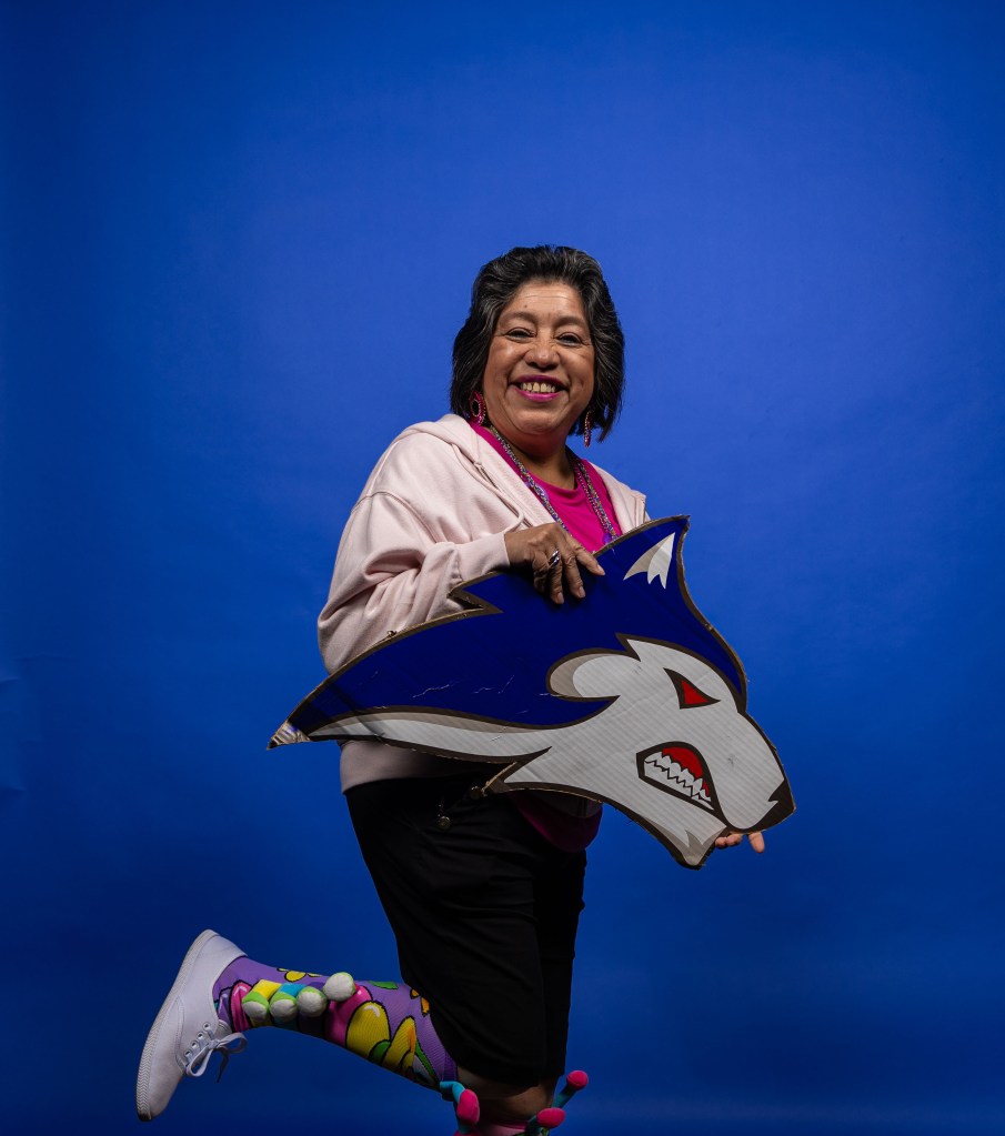 A joyful woman wearing a pink hoodie and colorful patterned leggings is playfully posing while holding a blue and white wolf mascot sign, set against a bright blue background.