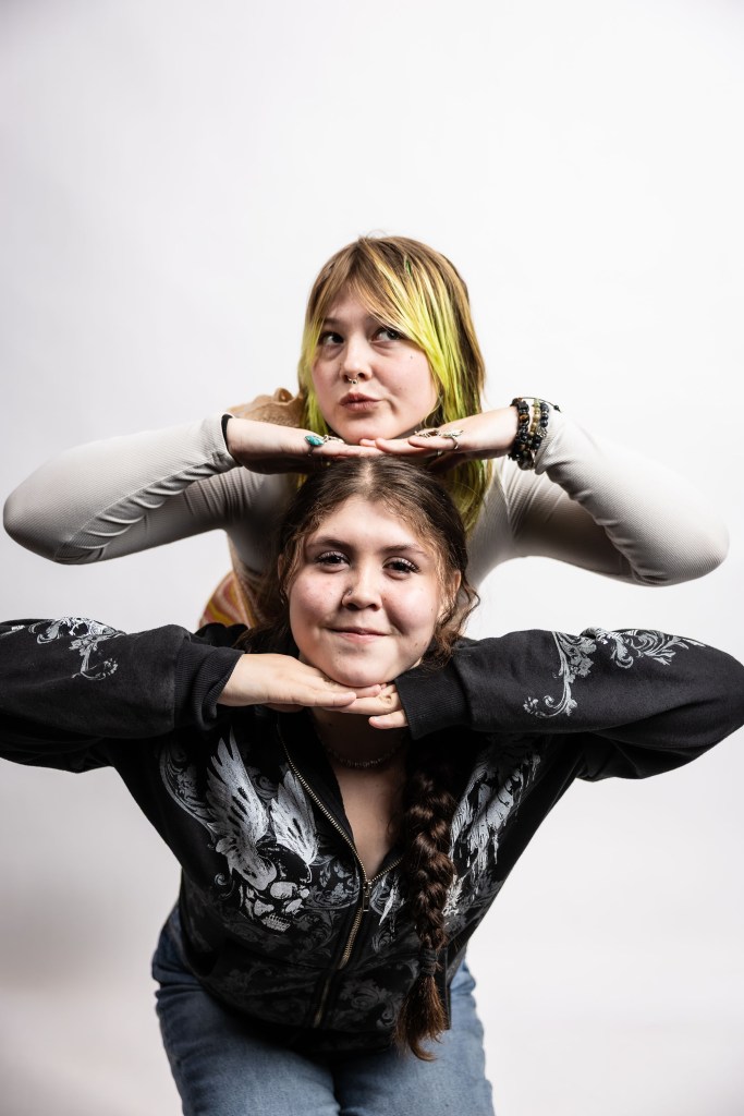 Two young women posing playfully, one standing behind the other with hands on their chin, against a plain white background.