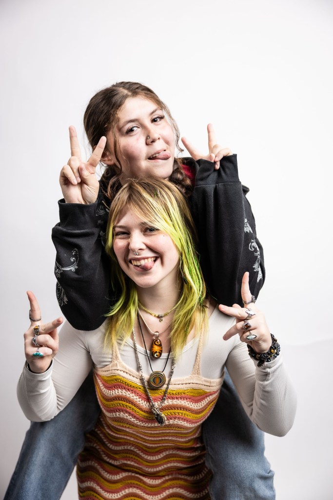 Two young women posing playfully for the camera, one sitting on the other's shoulders, both making peace signs with their fingers. The woman on top is sticking out her tongue, while the woman below smiles widely. They are wearing casual, colorful clothing and accessories.