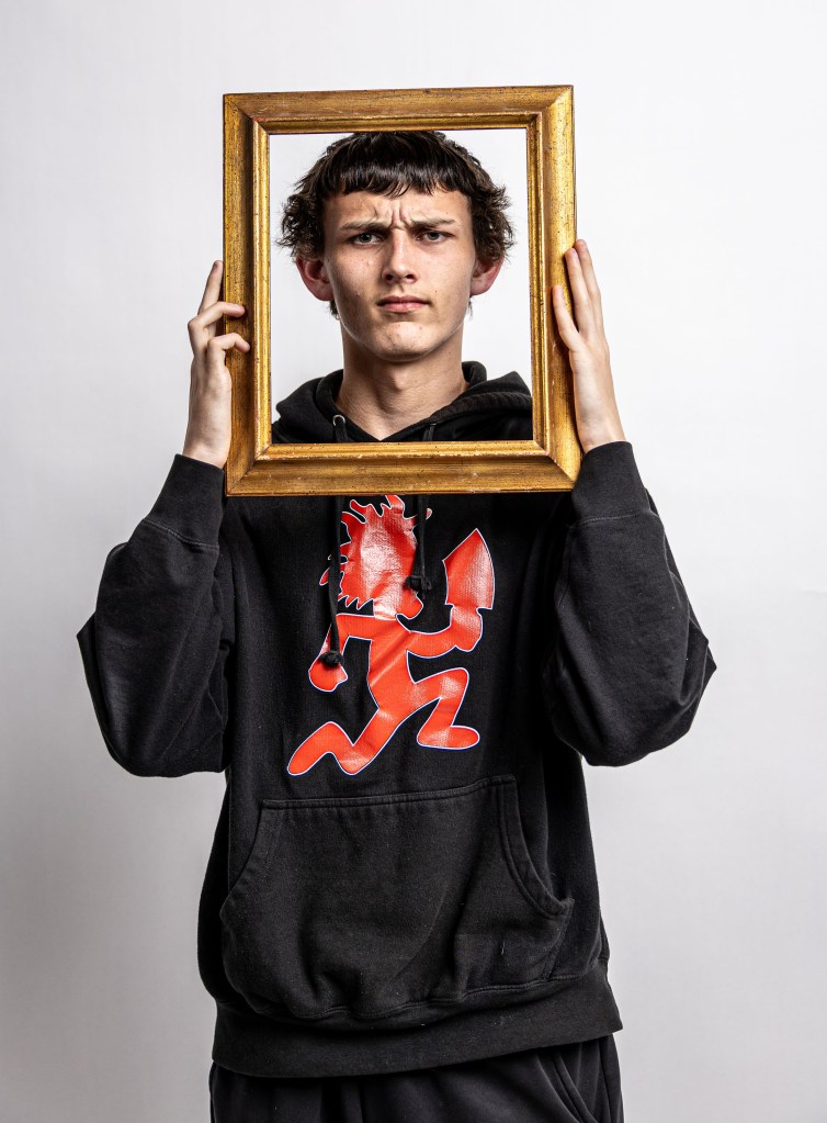 A young male holds a golden picture frame in front of his face, wearing a black hoodie with a red graphic design, against a plain white background.