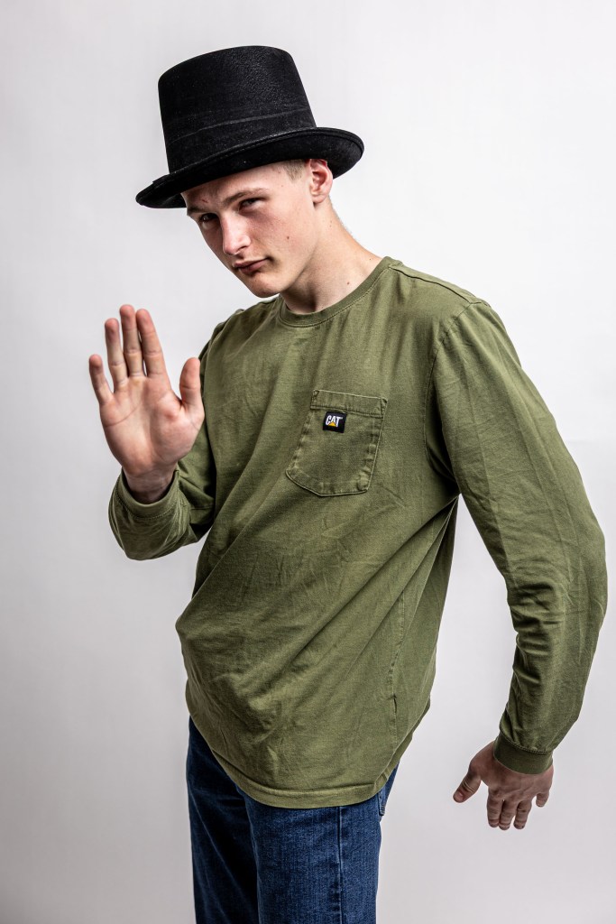A young man wearing a black top hat and an olive green long-sleeve shirt, standing with one hand raised in a casual gesture.