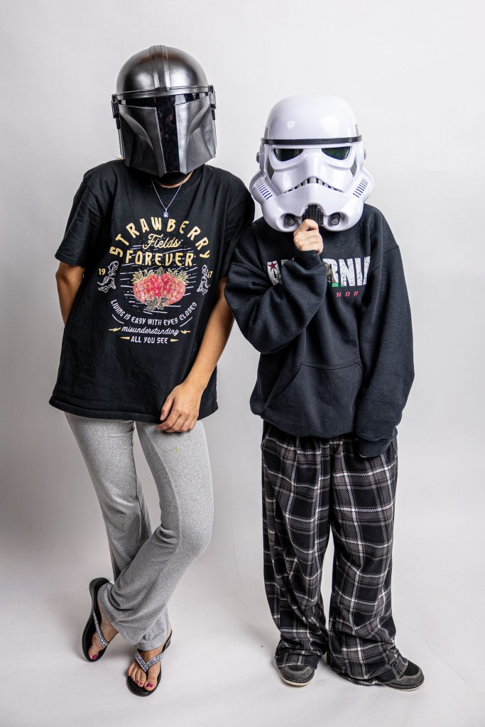 Two individuals wearing character masks, one in a black helmet and the other in a white stormtrooper mask. They are dressed casually, with one wearing a t-shirt and gray pants, and the other in a hoodie and plaid pajama pants, posing against a neutral backdrop.