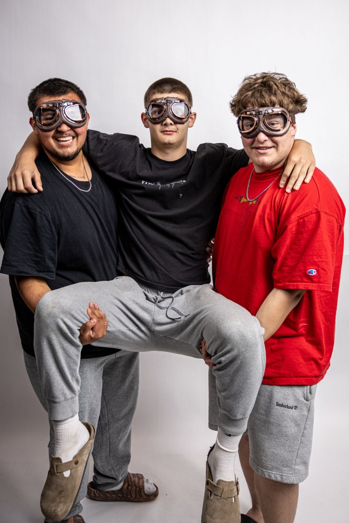 Three young men posing together, wearing goggles and casual clothing. One man is sitting on the other's lap, while the other two stand beside him with their arms around each other.
