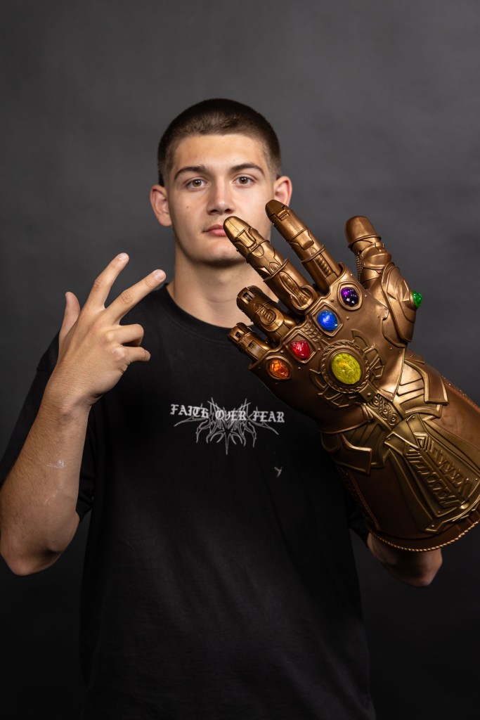 A young man wearing a black t-shirt poses with a large replica of the Infinity Gauntlet, featuring colorful gems, against a dark background.