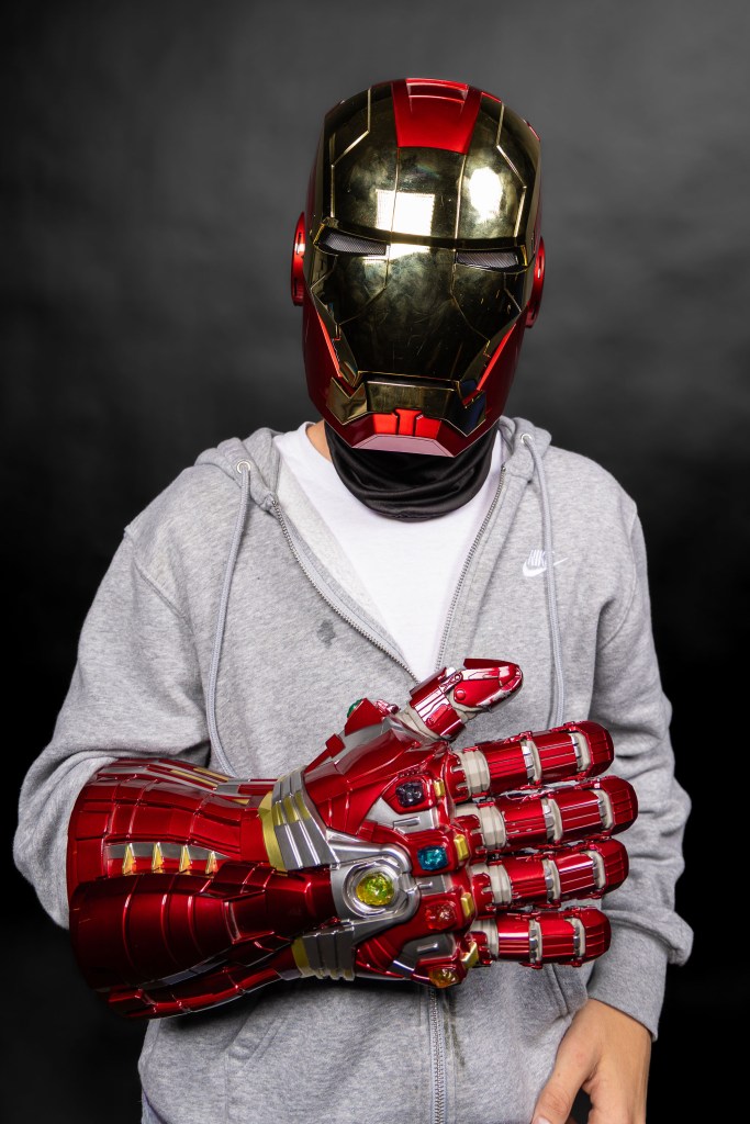 Person wearing an Iron Man helmet and a gray hoodie, holding a red and silver superhero gauntlet with jewel details, against a dark background.